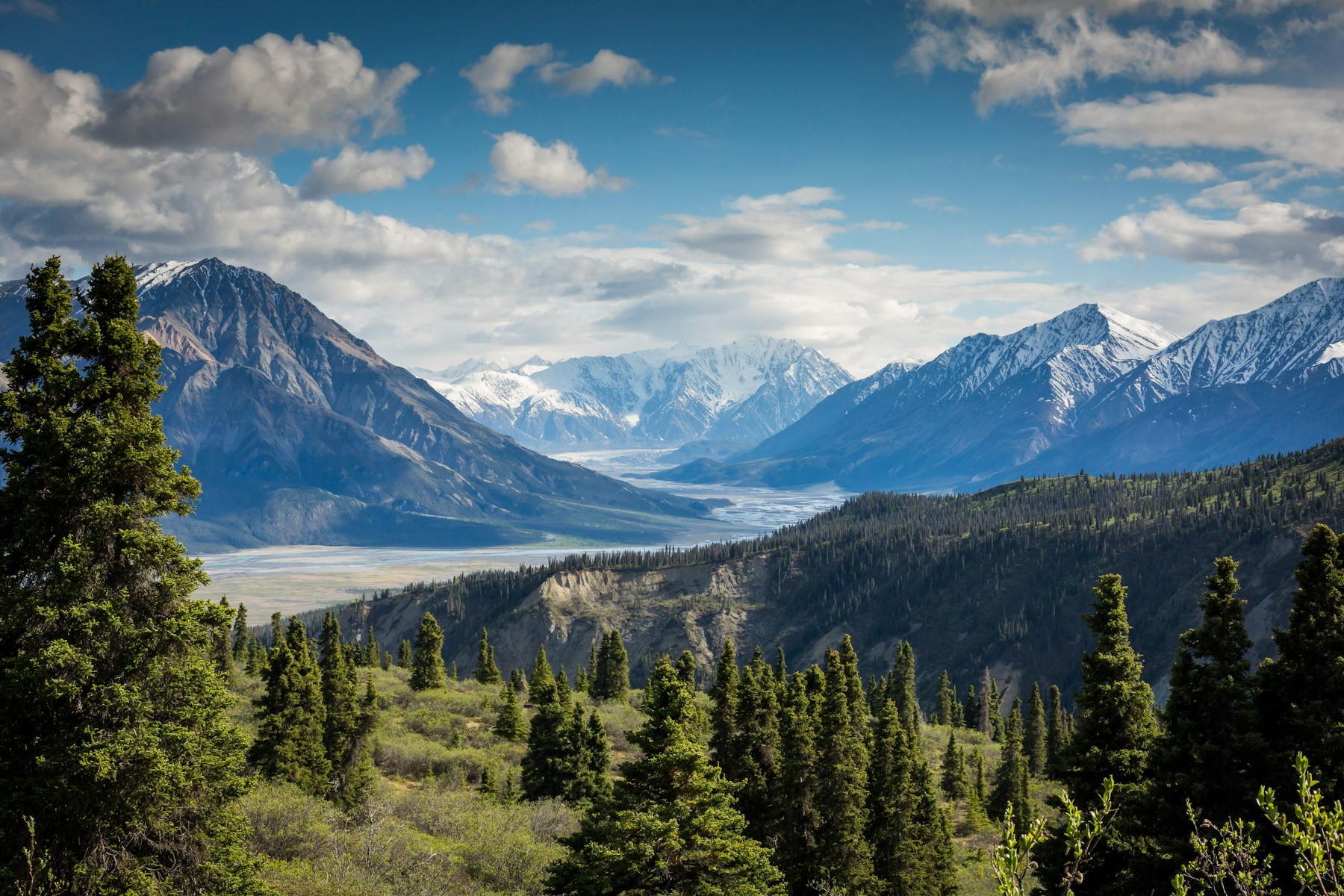A green river valley between snowy mountains under a cloud-filled sky